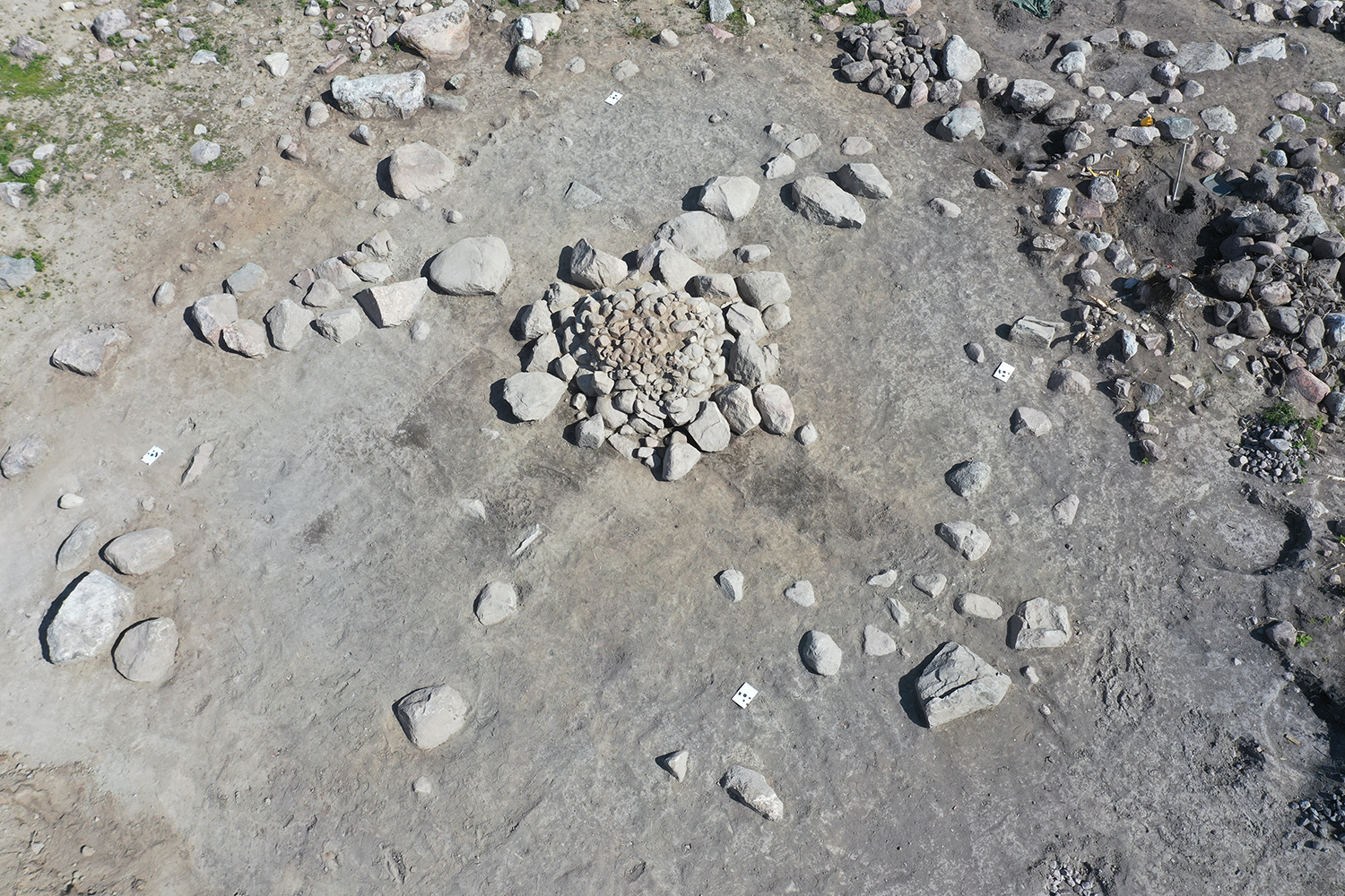 Jordmanteln dolde flera stenkonstruktioner. I mitten kärnröset omgivet av en krets av större stenar. I bildens övre del två närmast ovala stenformationer med än så länge okänd funktion. Möjligen har andra stenar bildat en kantkedja utmed högens yttre kanter. Drönarfoto: Arkeologerna.