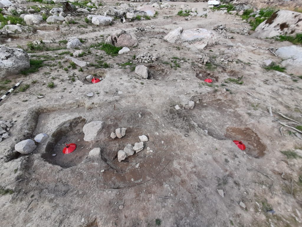 Nedgrävningar för takbärande stolpar i det mindre huset har markerats med röda koner. I förgrunden en grop med mindre stenar på rad, mellan stenarna fanns en mindre platta av bränd lera som kan ha fungerat som bakplatta i en eldstad.