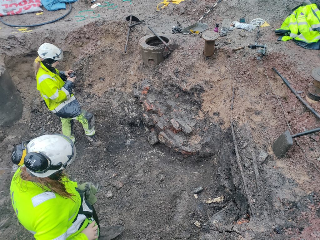 Två arkeologer dokumenterar en äldre tegelkonstruktion på marken,