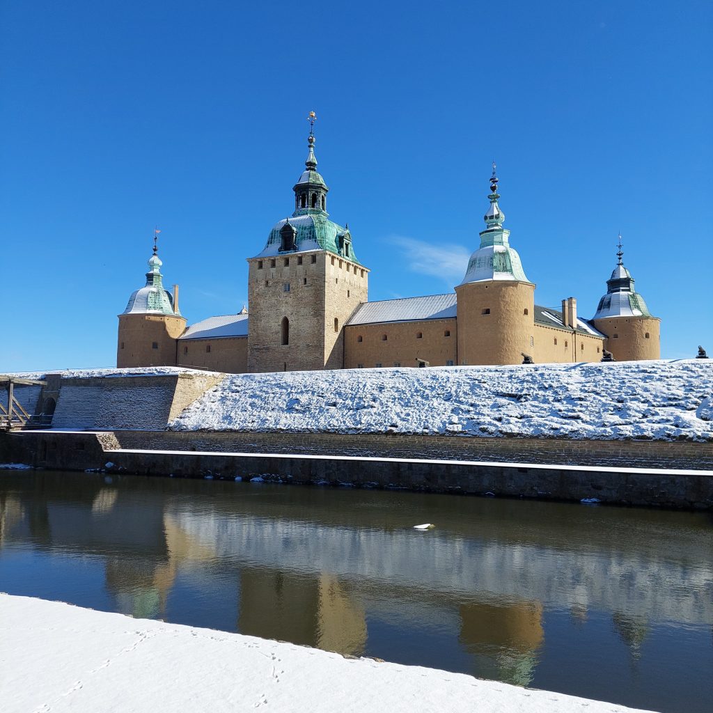Kalmar slott och vallgrav mot en blå himmel. Marken och vallen invid vallgraven är täckt av snö. 