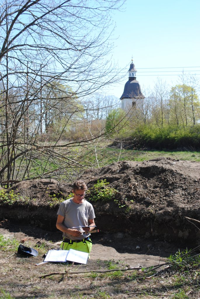 En person står i ett grävschakt, i bakgrunden syns en kyrka som sticker upp ur buskarna.