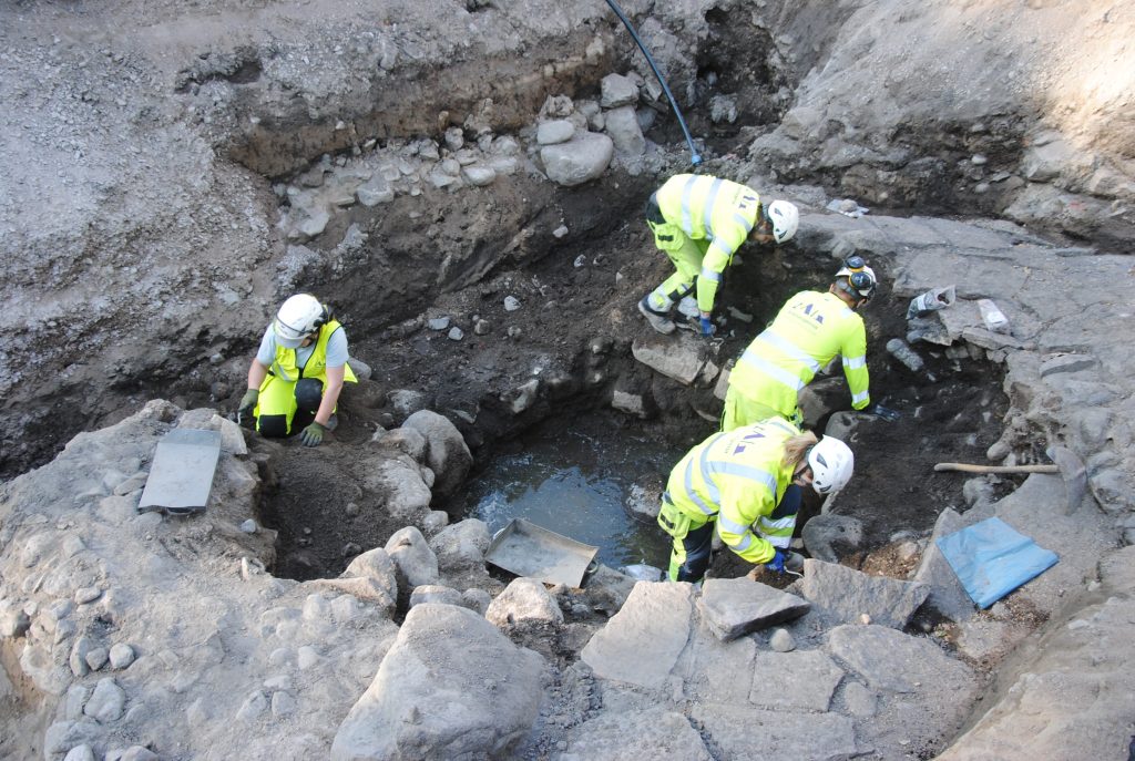 Brunnen undersöks av Frida, Adrian, Björn och Maja. På utsidan syns rester av kalkstensgolv från en äldre byggnad som förstörts i samband med att brunnen grävdes på 1500-talet.