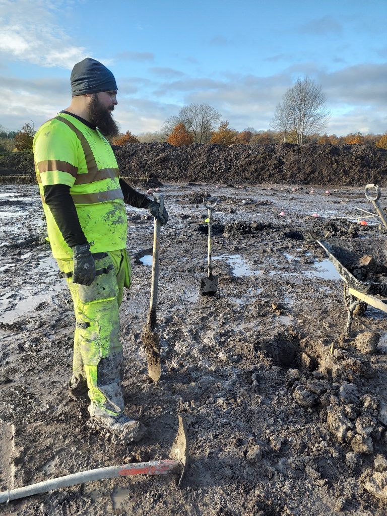 Arkeolog som står och stödjer sig på en spade och tittar ner på den leriga marken.