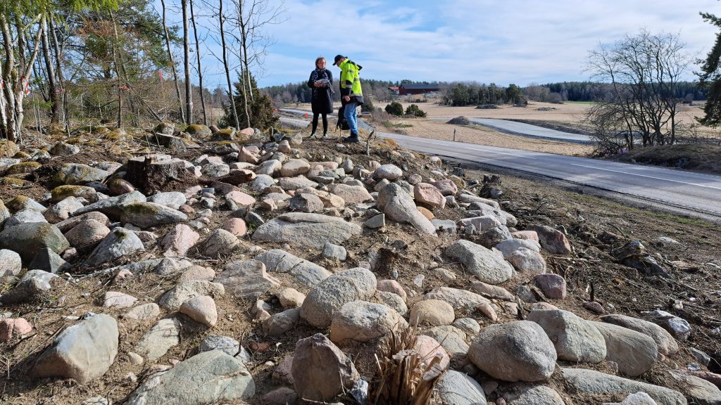 Två personer står ovanpå en stenhög, en stensättning, med vägen i bakgrunden.