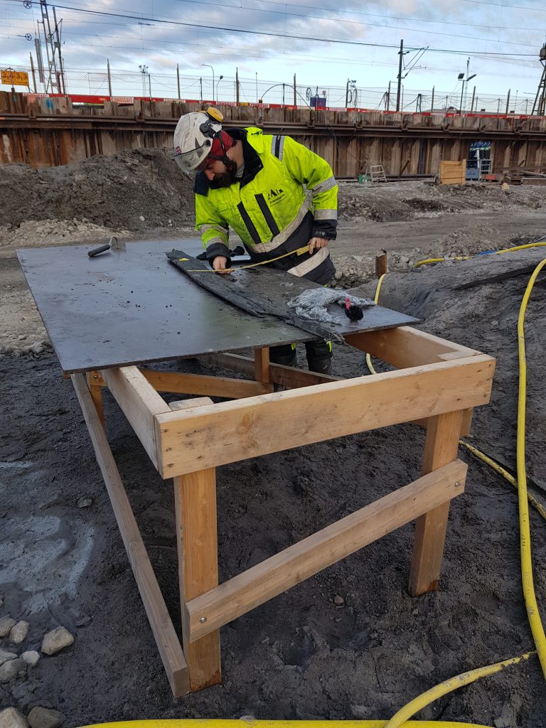 En person i gult står vid ett bord och mäter på en vrakdel av trä.