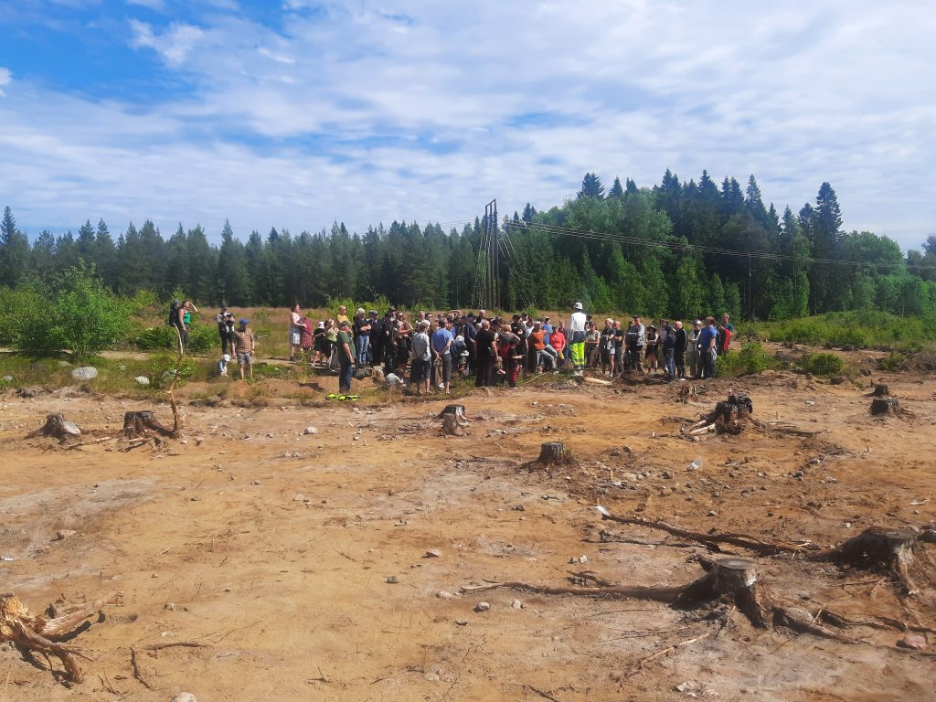 En stor grupp människor står på en öppen yta med skog bakom medan en person pratar och visar.