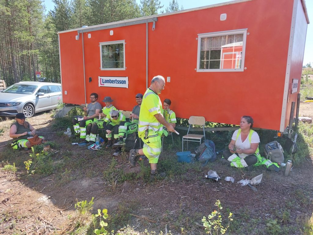 En arkeolog delar ut glassar till arkeologer som tar en paus i skuggan vid en röd arbetsbod.