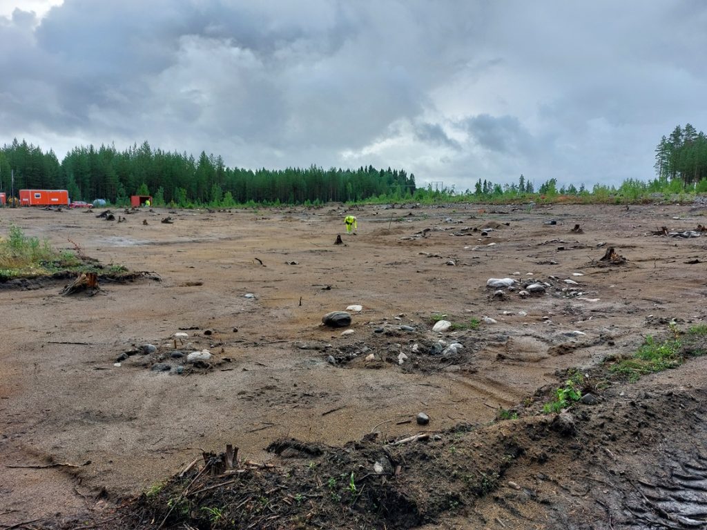 En bild på en tom plats med jord och stenar strödda, skogen i bakgrunden. 
