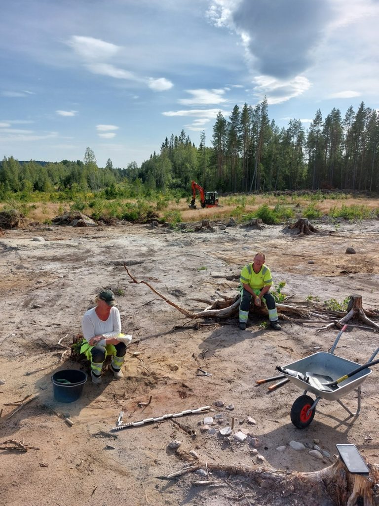 Två arkeologer sitter på ett öppet landskap, en skottkärra i förgrunden och skog i bakgrunden.