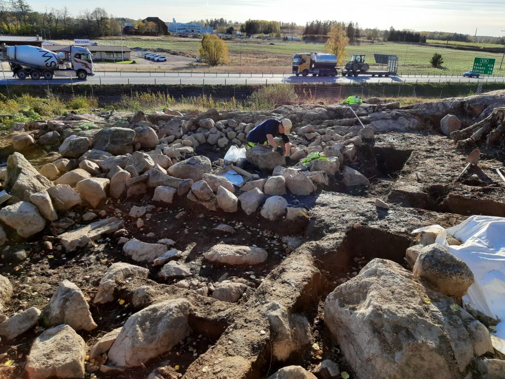 En arkeolog gräver ett område som är indelat i fyra delar, en väg går i bakgrunden.