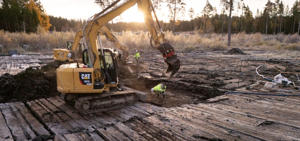 En grävmaskin undersöker en våtmark, skog i bakgrunden.