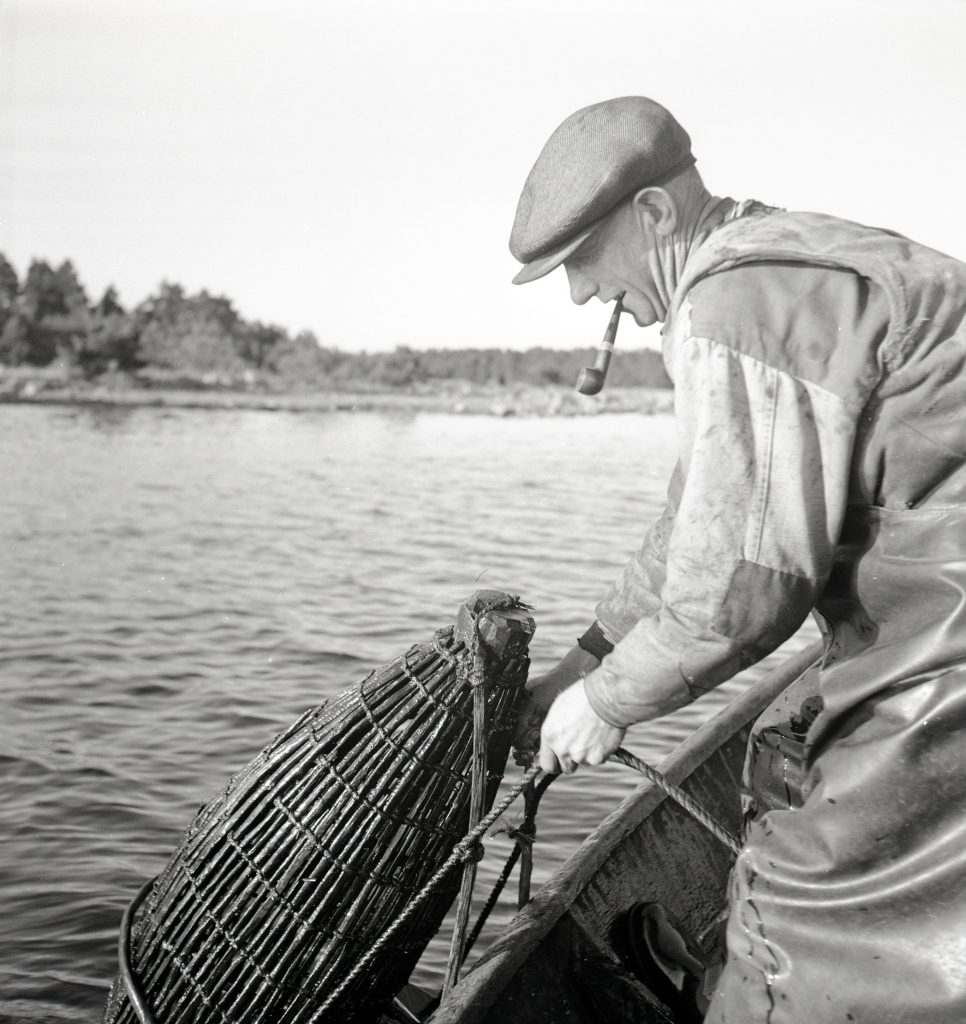 Ett svartvitt foto på en man som vittjar en mjärde med en sjö i bakgrunden.