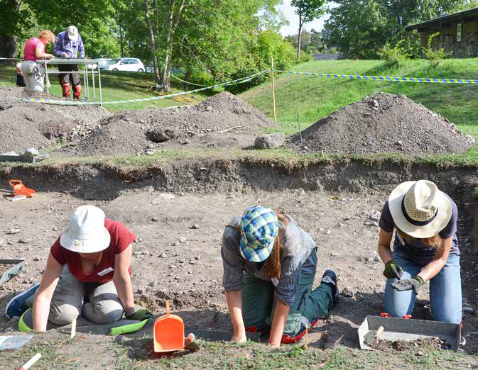 Tre personer gräver, i bakgrunden syns kullar, jordhögar och ett såll där två andra personer jobbar.