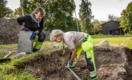 Två arkeologer kvinnliga håller på och gräver i gräsmattan, en grop.