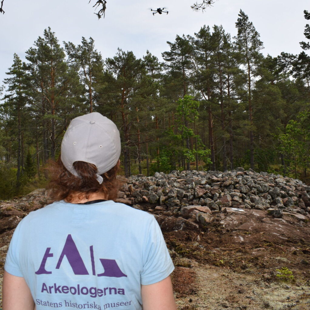 En bild på en kvinna i ljusblå t-shirt med Arkeologernas logga som flyger en drönare.