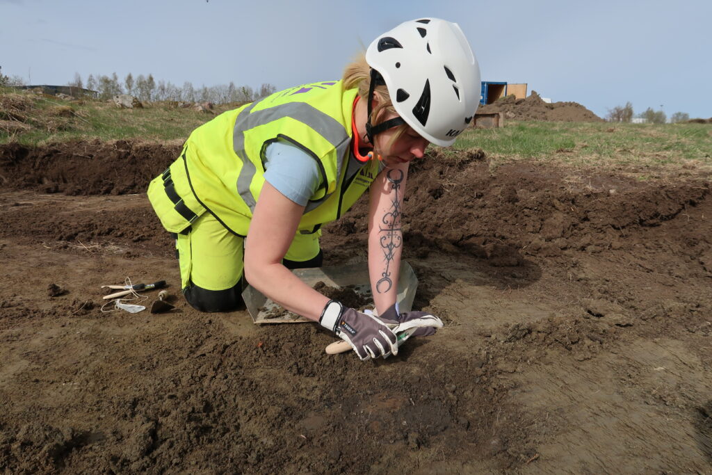 En arkeolog ligger på marken iförd vit hjälm och gräver i jorden.