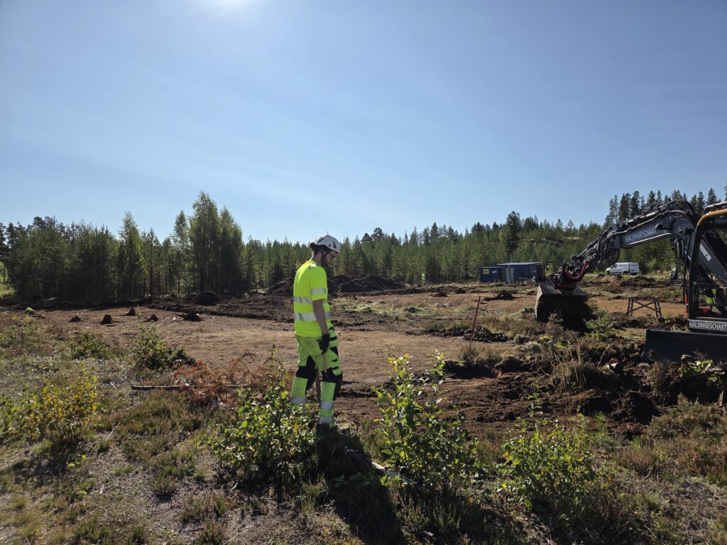 Arkeollog ute på öppen mark med grävskopa till höger, i förgrunden låga buskar och i bakgrunden skog. 
