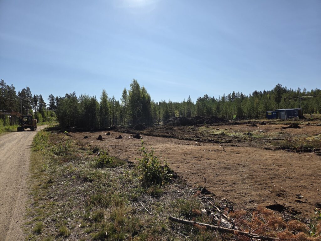 En vidsträckt plats, avbanad jordyta med barrskog bakom. Solen lyser från en blå himmel.
