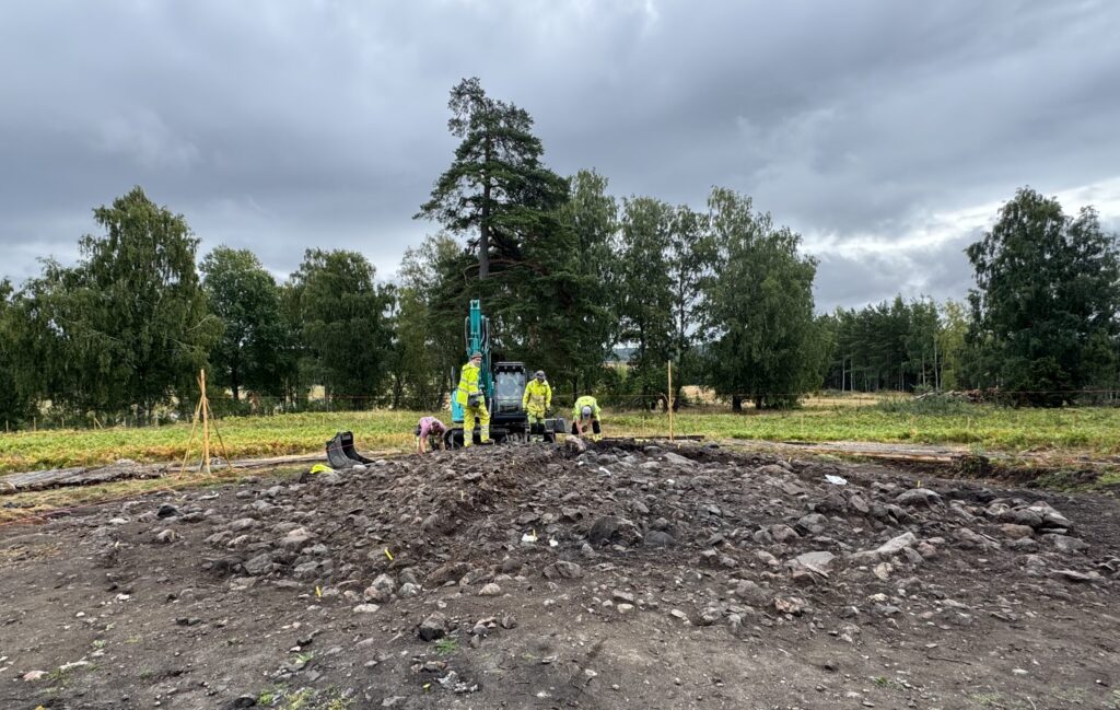 Tre personer och en grävskopa som befinner sig uppe på en hög med stenar, i bakgrunden gräs och träd. 