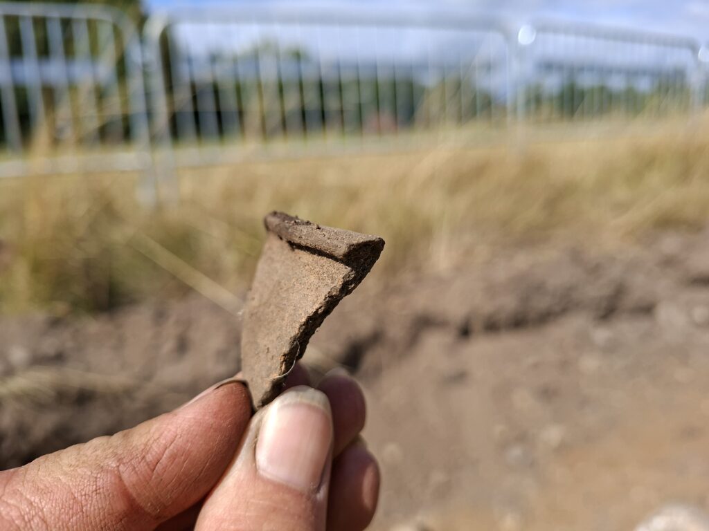 En liten keramikskärva, en mynningsbit hålls fram av några fingrar, i bakgrunden en äng och åker. 