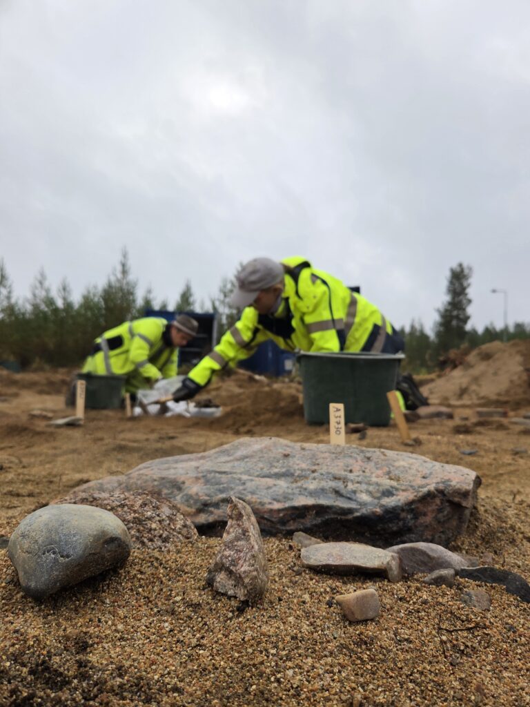 Två arkeologer gräver med skog och ljus himmel bakom. I förgrunden stenar och gula markeringspinnar mot en brun-beige mark.